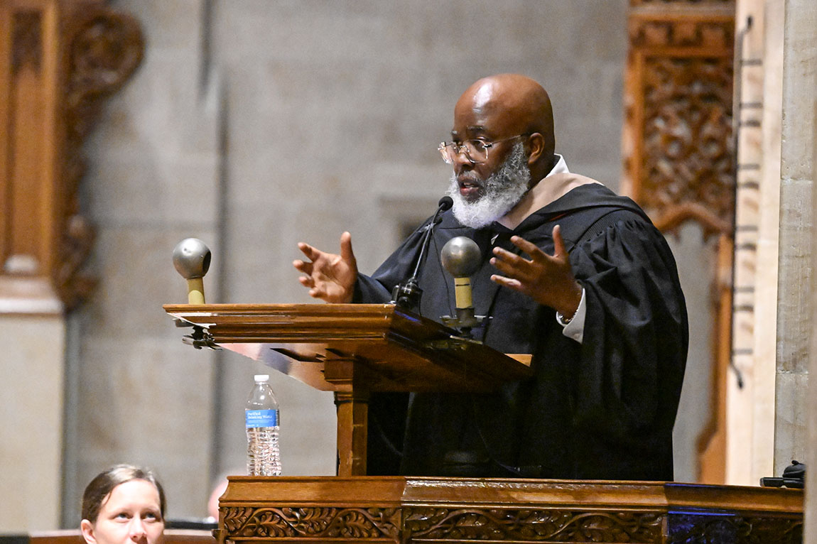 A bald man with a gray beard and glasses speaks at a podium